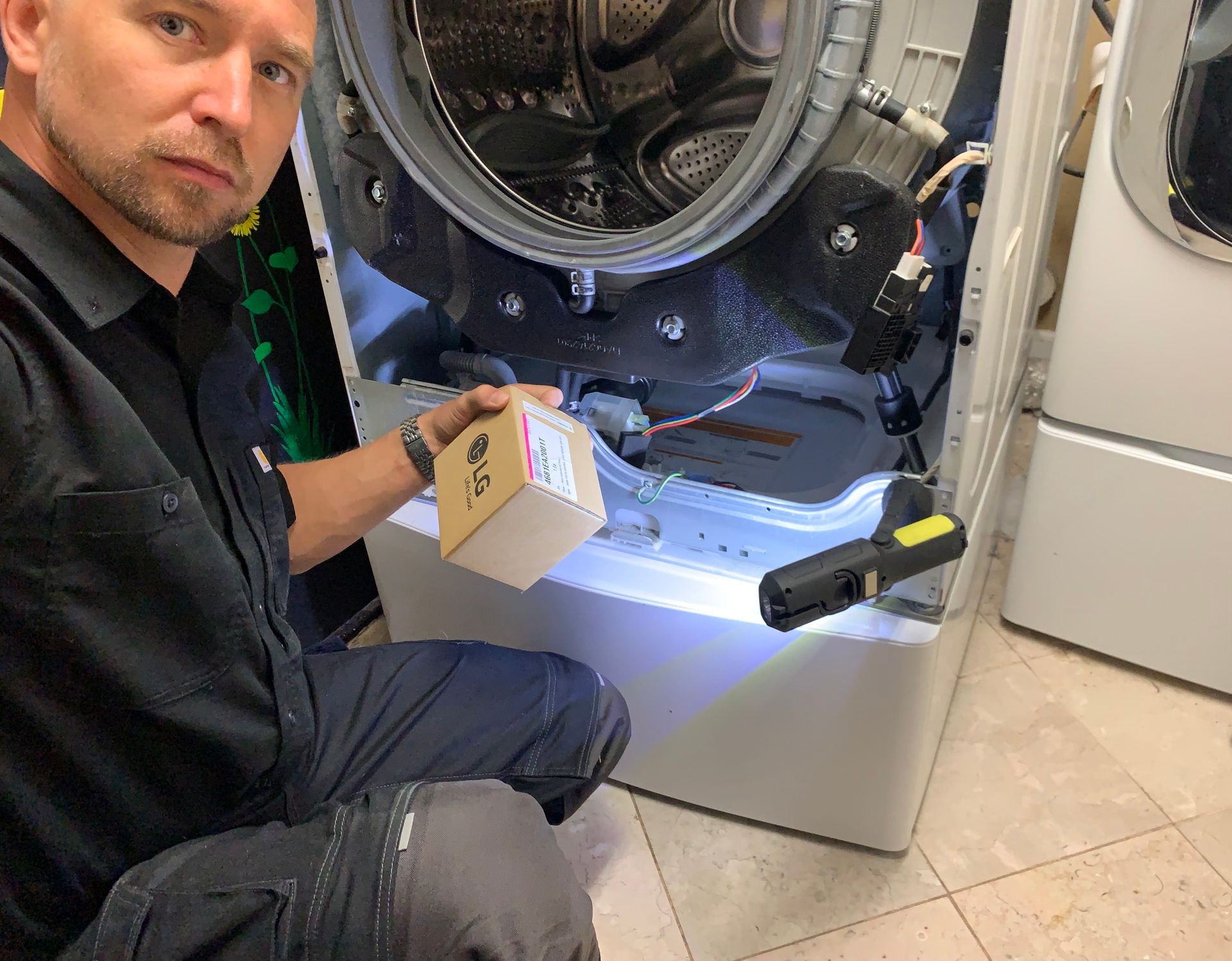 Person repairing a washing machine, holding a replacement part and flashlight, with front panel removed.
