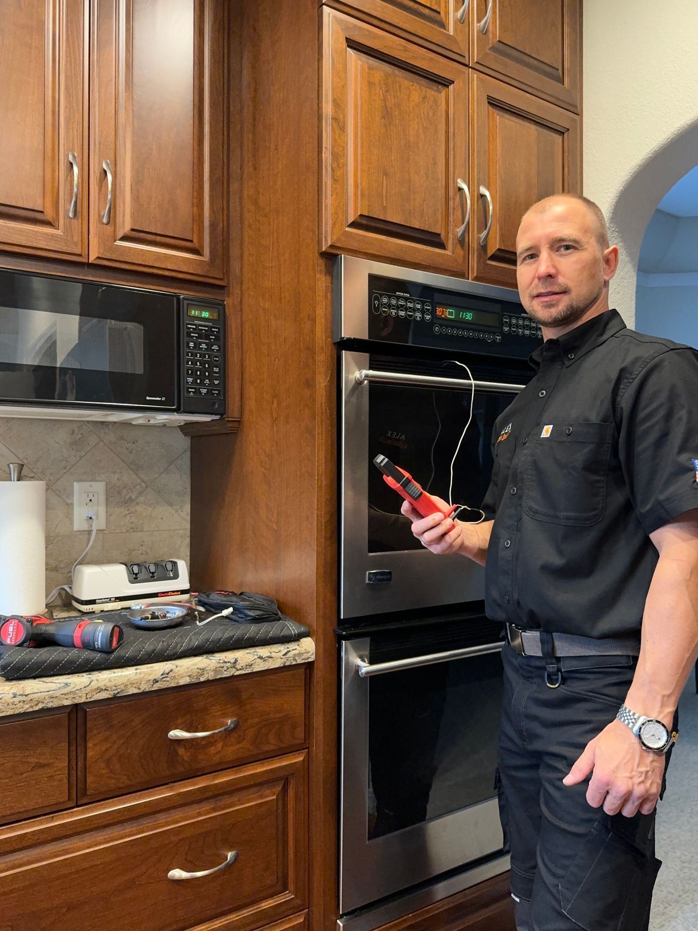 Man in black uniform holding equipment next to a kitchen oven, surrounded by wooden cabinets.